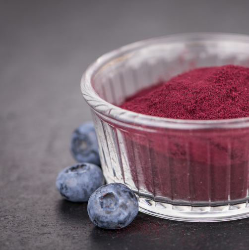 Deep purple blueberry powder in a clear glass bowl beside fresh blueberries