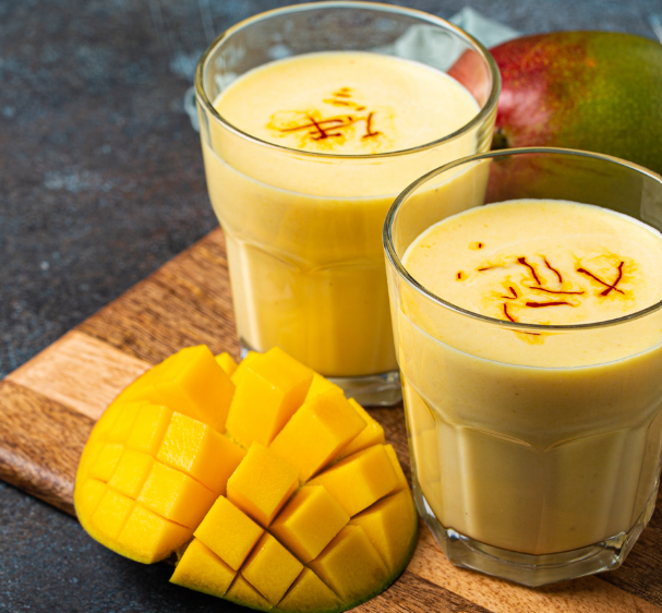 Two glasses of creamy yellow mango drink beside freshly cut mango on a wooden board